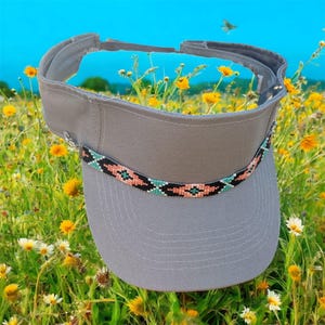 May include: A gray visor with a beaded band featuring a geometric pattern in black, coral, and turquoise. The visor is set against a backdrop of a field of yellow wildflowers and a blue sky.