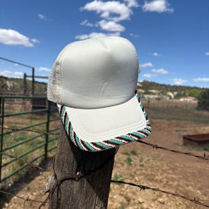 May include: A white trucker cap featuring a mesh back and a structured front panel. The brim is decorated with a woven pattern of diagonal stripes in alternating colours of turquoise, black, and white, creating a distinct geometric border design.