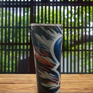 Puede incluir: Un vaso alto y aislado con un diseño patriótico. El vaso presenta un patrón abstracto azul, blanco y rojo, con una vista parcial de un círculo azul. El fondo es una vista borrosa de una superficie de madera y una ventana.