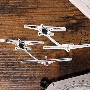 Può includere: Quattro adesivi di aerei su una superficie di legno. Gli adesivi raffigurano vari modelli di aeromobili, tra cui un aliante e piccoli aerei. Gli adesivi sono bianchi con dettagli neri e rossi. Uno strumento di navigazione e una mappa sono parzialmente visibili sullo sfondo.