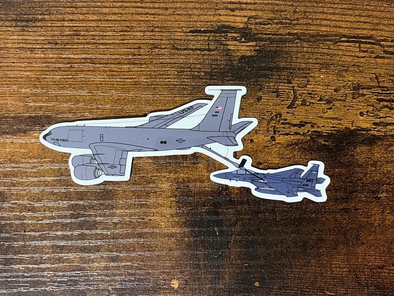 May include: A sticker depicting two military aircraft. The larger aircraft is a grey tanker with "U.S. AIR FORCE" printed on the side. The smaller aircraft is a grey fighter jet, connected to the tanker by a refueling boom.