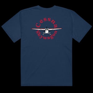 Puede incluir: Una camiseta azul marino con un gráfico circular rojo en la espalda. El gráfico presenta un avión pequeño con el texto "Cessna Skywagon" en una fuente retro roja.