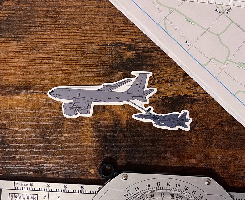 May include: A sticker featuring a gray and white military aircraft, a Boeing KC-135 Stratotanker, refueling a gray and white F-15 Eagle fighter jet.