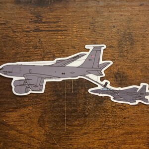 May include: A sticker depicting a gray military aircraft and a smaller jet, linked by a refueling line. The larger aircraft has the text "U.S. Air Force" and "USAF". The sticker is on a wooden surface.