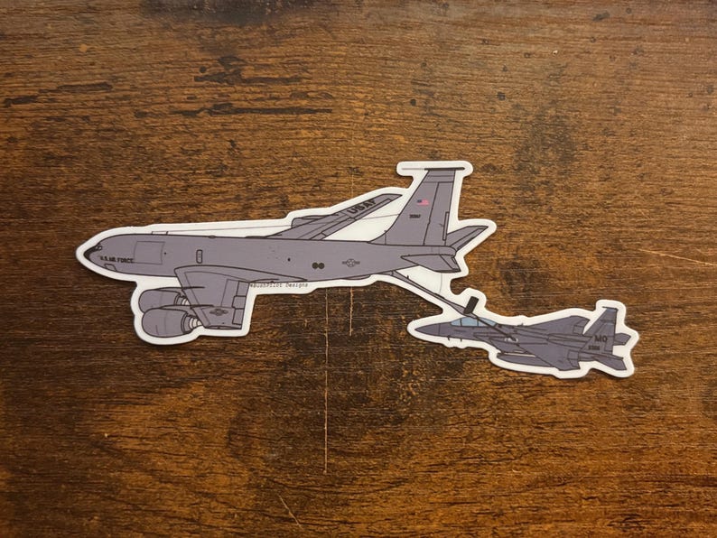May include: A sticker depicting a gray military aircraft and a smaller jet, linked by a refueling line. The larger aircraft has the text "U.S. Air Force" and "USAF". The sticker is on a wooden surface.