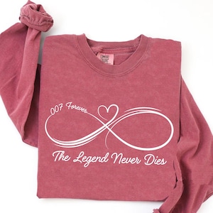 May include: A maroon long-sleeve shirt with the text "007 Forever" and "The Legend Never Dies" in white. The design includes an infinity symbol with a heart. The shirt is made of a soft material.