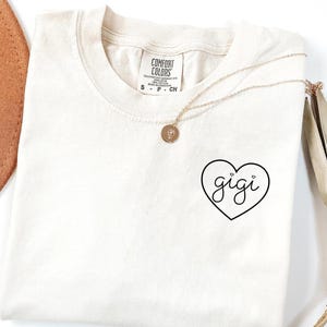 Op de afbeelding: Een ivoorwit T-shirt met een zwart hartontwerp met daarin "gigi". Het hart heeft twee kleine hartjes erboven.
