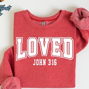 May include: A heather scarlet red sweatshirt with the word "LOVED" in white block letters, arched above "JOHN 3:16". The sweatshirt has a crew neck and folded sleeves. The text "HEATHER SCARLET RED" is printed at the bottom.