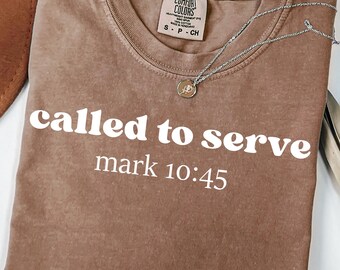 May include: Espresso-colored t-shirt with the text "called to serve mark 10:45" in white. The shirt has a crew neck and short sleeves. A silver necklace with a pendant is draped across the shirt.