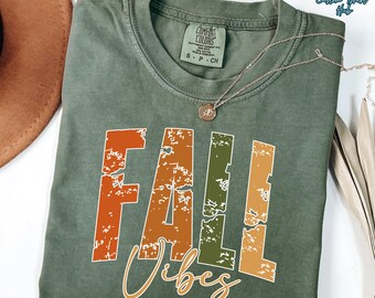 May include: Moss green t-shirt with the words "FALL Vibes" in orange, green, and tan distressed lettering. A silver necklace with a pendant is draped across the shirt. The shirt is folded on a surface with a brown hat.