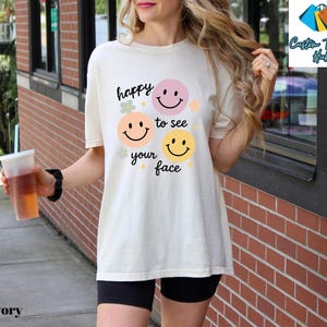 May include: Ivory-colored t-shirt with the text "happy to see your face" and smiley face graphics in pink, peach, and yellow. The shirt is a relaxed fit and is paired with black shorts. The image also shows a clear plastic cup with a straw.