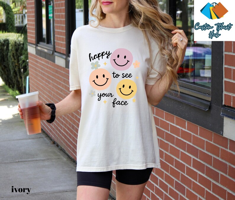 May include: Ivory-colored t-shirt with the text "happy to see your face" and smiley face graphics in pink, peach, and yellow. The shirt is a relaxed fit and is paired with black shorts. The image also shows a clear plastic cup with a straw.