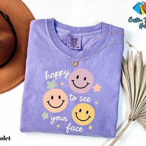 May include: A violet Comfort Colors t-shirt with a graphic of three smiley faces and the text "happy to see your face". The shirt has a crew neck and short sleeves. A gold necklace is draped across the shirt.