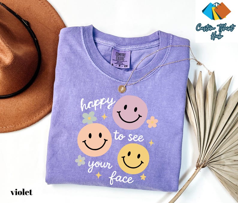 May include: A violet Comfort Colors t-shirt with a graphic of three smiley faces and the text "happy to see your face". The shirt has a crew neck and short sleeves. A gold necklace is draped across the shirt.