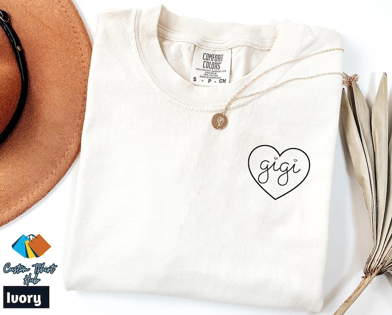Op de afbeelding: Een ivoorwit T-shirt met een zwart hartontwerp met daarin "gigi". Het hart heeft twee kleine hartjes erboven.