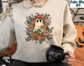 May include: A beige crewneck sweatshirt featuring a central graphic of a ghost wearing a brown hat, surrounded by orange, yellow, and green flowers and foliage. The ghost is positioned above a small pumpkin. The sweatshirt has long sleeves.