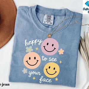 May include: A blue jean colored t-shirt with the phrase "happy to see your face" and three smiley faces in pink, peach, and yellow. The shirt has a crew neck and short sleeves. A gold necklace is draped across the shirt.