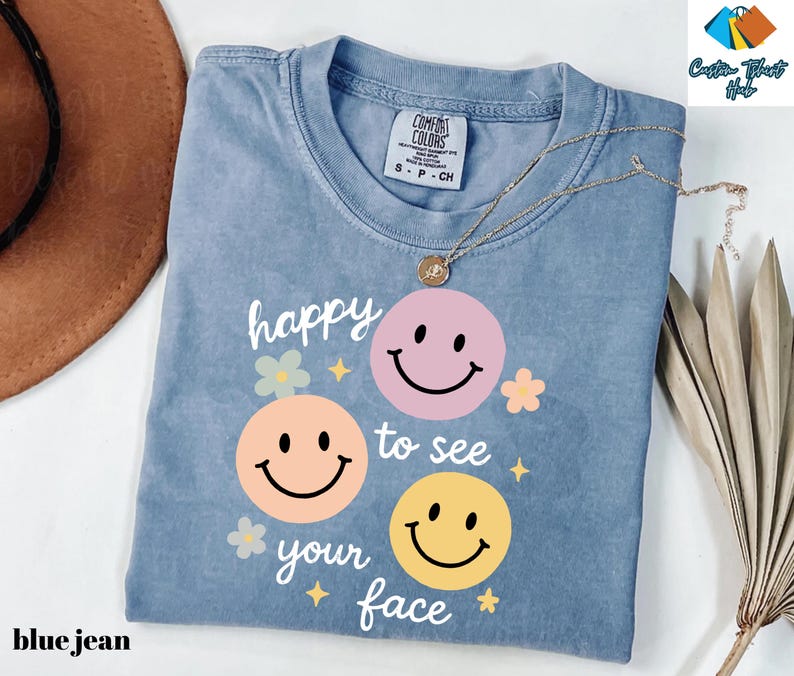 May include: A blue jean colored t-shirt with the phrase "happy to see your face" and three smiley faces in pink, peach, and yellow. The shirt has a crew neck and short sleeves. A gold necklace is draped across the shirt.