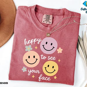 May include: Crimson-colored t-shirt with the phrase "happy to see your face" and three smiley face graphics in pink, peach, and yellow. The shirt has a crew neck and a Comfort Colors tag. A silver necklace with a pendant is on the shirt.