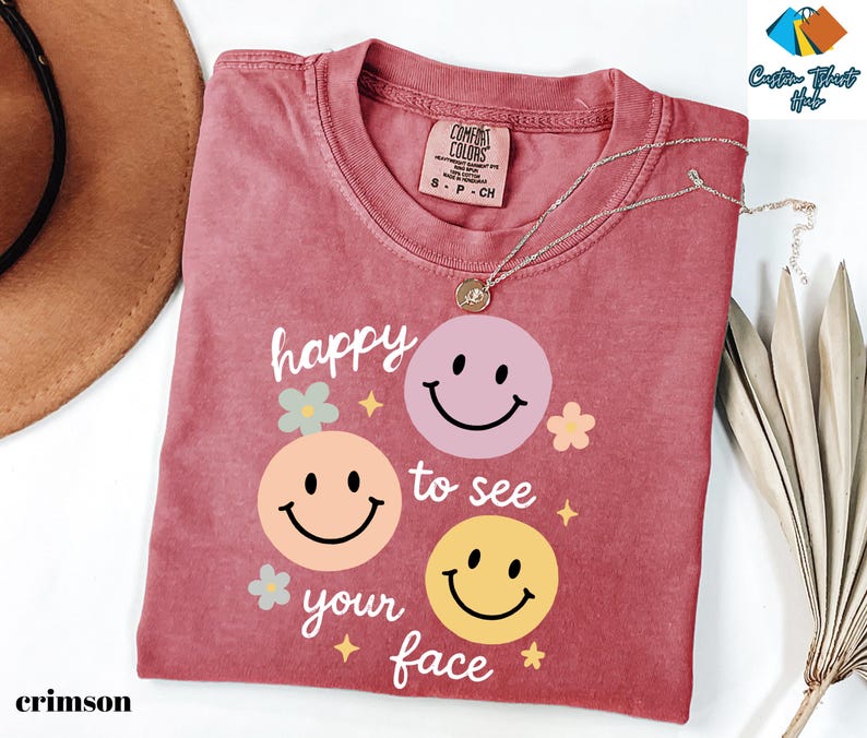May include: Crimson-colored t-shirt with the phrase "happy to see your face" and three smiley face graphics in pink, peach, and yellow. The shirt has a crew neck and a Comfort Colors tag. A silver necklace with a pendant is on the shirt.