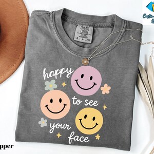 May include: A gray t-shirt with the phrase "happy to see your face" and three smiley face graphics in pink, peach, and yellow. The shirt has a crew neck and short sleeves. A silver necklace with a pendant is on the shirt.