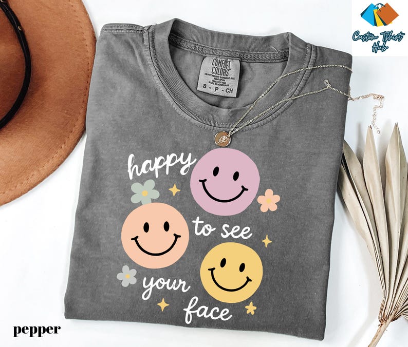 May include: A gray t-shirt with the phrase "happy to see your face" and three smiley face graphics in pink, peach, and yellow. The shirt has a crew neck and short sleeves. A silver necklace with a pendant is on the shirt.
