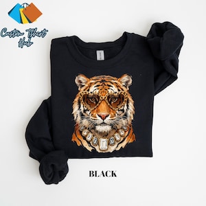 May include: A black crewneck sweatshirt featuring a graphic illustration of a tiger wearing heart-shaped leopard print sunglasses and a chunky jewelled necklace with large clear stones. The word BLACK is printed in simple black text below the tiger.