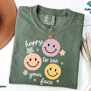 May include: Moss green t-shirt with a graphic of three smiley faces in pink, peach, and yellow. The text "happy to see your face" is printed below the faces. A gold necklace is draped over the shirt.