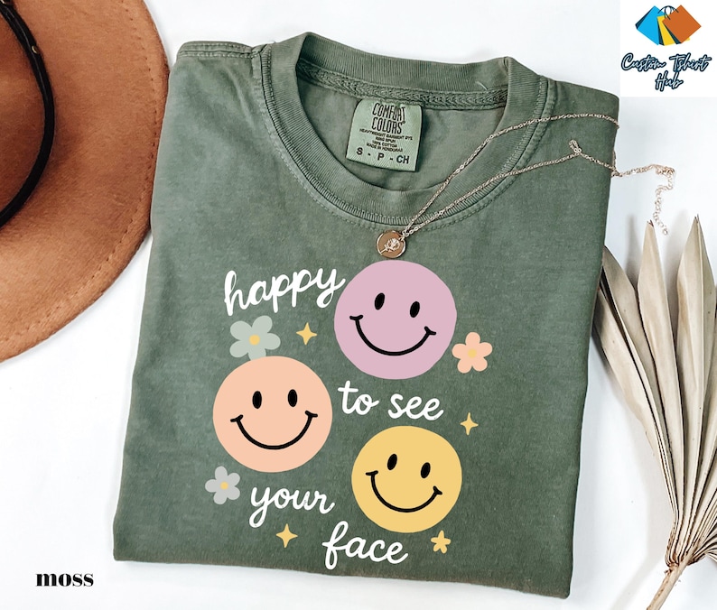 May include: Moss green t-shirt with a graphic of three smiley faces in pink, peach, and yellow. The text "happy to see your face" is printed below the faces. A gold necklace is draped over the shirt.