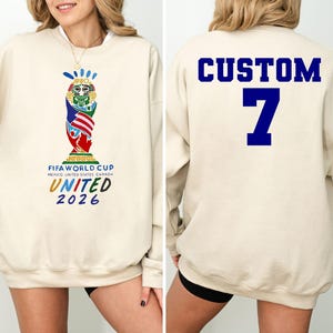 May include: Beige sweatshirt featuring the FIFA World Cup United 2026 logo on the front. The back displays the word "CUSTOM" above the number "7" in blue. The sweatshirt is a long-sleeved, casual garment.