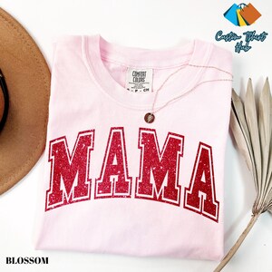 May include: A light pink Comfort Colors t-shirt with the word "MAMA" in large, red, glittery block letters. A delicate necklace with a pendant rests on the shirt. The word "BLOSSOM" is printed in black at the bottom left.