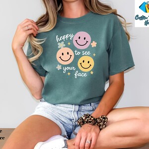 May include: A blue spruce t-shirt with the phrase "happy to see your face" and three smiley face graphics in pink, orange, and yellow. The shirt has a relaxed fit and is paired with denim shorts.