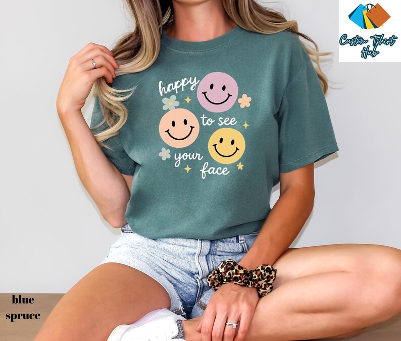 May include: A blue spruce t-shirt with the phrase "happy to see your face" and three smiley face graphics in pink, orange, and yellow. The shirt has a relaxed fit and is paired with denim shorts.