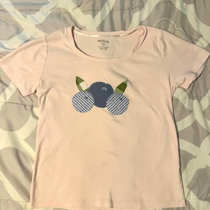 May include: A light pink t-shirt with a crew neck. The shirt features an appliqué design of three blueberries, with two checkered blue and white berries and one solid blue berry. Green leaves are on either side of the berries. The brand name is visible on the tag.