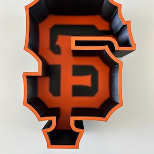 May include: A 3D printed, black and orange, hollow, SF logo. The logo is a stylized letter "S" with a smaller "F" inside.