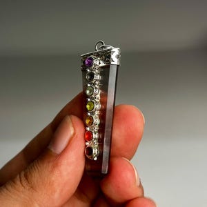 May include: A silver-toned pendant featuring a smoky quartz crystal with seven colorful gemstones. The gemstones are arranged vertically, representing the chakras. The pendant is held in a hand against a gray background.