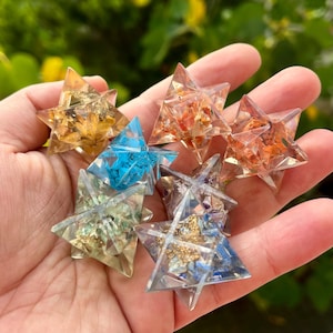 May include: A collection of clear, star-shaped resin ornaments, each filled with colorful dried flowers and crystals. The ornaments are in shades of yellow, orange, blue, and clear, creating a vibrant and eye-catching display.