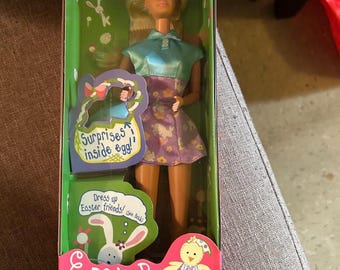 May include: A boxed Easter Surprise Barbie doll. The doll has blonde hair and wears a blue top and a purple floral skirt. The packaging features Easter-themed graphics, including a bunny and chick, with the text "Easter Surprise Barbie".
