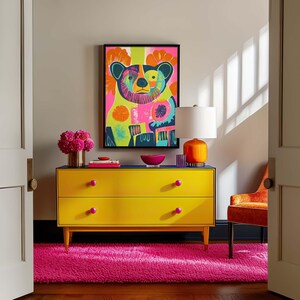 Op de afbeelding: Een gele commode met twee laden en roze knoppen staat op een roze shagtapijt. Een ingelijst afdruk van een kleurrijke beer hangt boven de commode. Een lamp met een witte kap en een oranje voet staat op de commode.