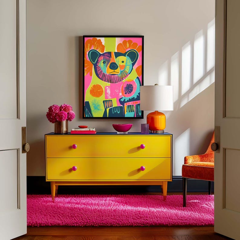 Op de afbeelding: Een gele commode met twee laden en roze knoppen staat op een roze shagtapijt. Een ingelijst afdruk van een kleurrijke beer hangt boven de commode. Een lamp met een witte kap en een oranje voet staat op de commode.