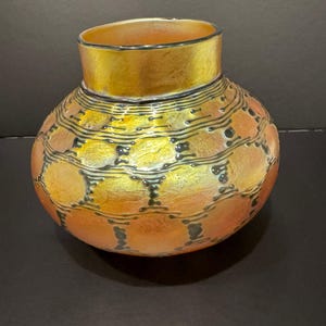 May include: A decorative glass vase with a wide, rounded body and a short, cylindrical neck. The vase features a metallic gold and orange color scheme with a pattern of black dots and lines. The rim is a darker gold.