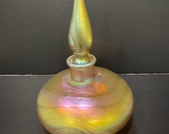 Puede incluir: Un frasco de perfume de vidrio iridiscente con un cuerpo bulboso y un tapón largo y puntiagudo. El frasco presenta un patrón arremolinado de tonos dorados, rosados y verdes. El tapón, de forma y color similares, realza el diseño elegante.