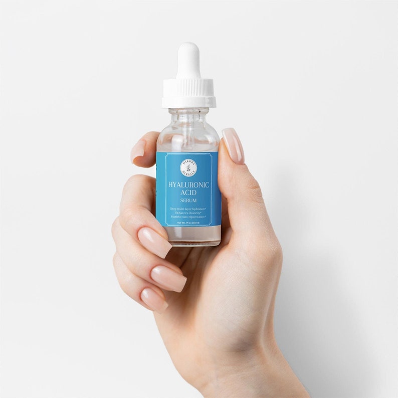Puede incluir: Una botella de vidrio transparente con una etiqueta azul que dice "HYALURONIC ACID SERUM". La botella tiene un gotero blanco y se sostiene en una mano.