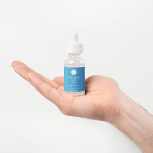 Puede incluir: Una botella de vidrio transparente con un gotero blanco y una etiqueta azul que dice "HYALURONIC ACID SERUM".