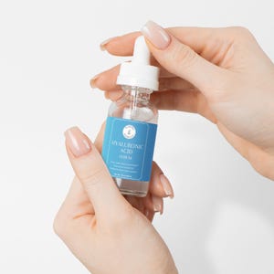 Puede incluir: Una botella de vidrio transparente con una etiqueta azul que dice "HYALURONIC ACID SERUM" y un tap&oacute;n cuentagotas blanco. La botella est&aacute; siendo sostenida por una mano.