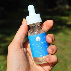 Puede incluir: Una botella de vidrio transparente con un gotero blanco y una etiqueta azul que dice "Hyaluronic Acid Serum" con el texto "Hidrata y humecta profundamente* Mejora la elasticidad de la piel* Rejuvenecimiento de la piel* Contenido neto: 30 ml (1 fl oz)"