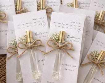 May include: A collection of wedding favors featuring small clear glass perfume vials with gold caps, tied with twine and baby's breath flowers. Each vial is attached to a white card printed with an August 2024 calendar, the text "Nuestro amor es eterno", and "Ester & Eloi 24.08.24".