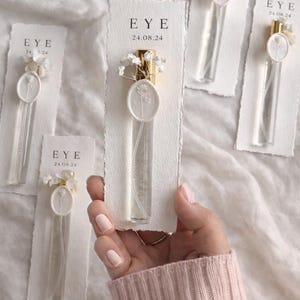 Könnte beinhalten: Klare Glasparfümflaschen mit goldenen Kappen und weißen Blumenakzenten werden auf weißen Karten präsentiert. Auf den Karten steht das Wort "EYE" und das Datum "24.08.24". Eine Person mit einem rosa Pullover hält die Flaschen.