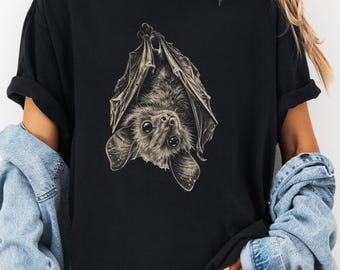 May include: A black short-sleeved t-shirt featuring a detailed, monochromatic illustration of a bat hanging upside down with its wings partially wrapped around its body. The bat has large, expressive eyes and textured fur, centered on the chest.