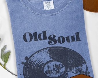 May include: A blue jean colored t-shirt with the words "Old Soul" and a graphic of a vinyl record. The shirt is a Comfort Colors brand, size L-G-G. A brown hat and greenery are in the background.